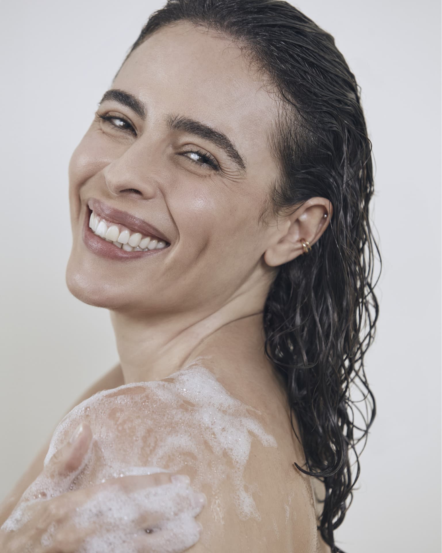 Mulher branca sorrindo com o cabelo longo molhado e ombros com espuma de banho