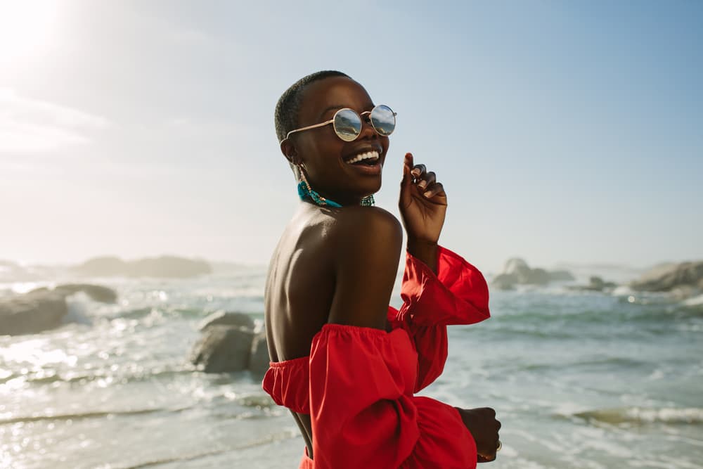 Imagem de mulher negra sorridente na praia com óculos de sol, brincos grandes na cor azul e uma blusa vermelha. Ao fundo dela há o mar e o céu azul