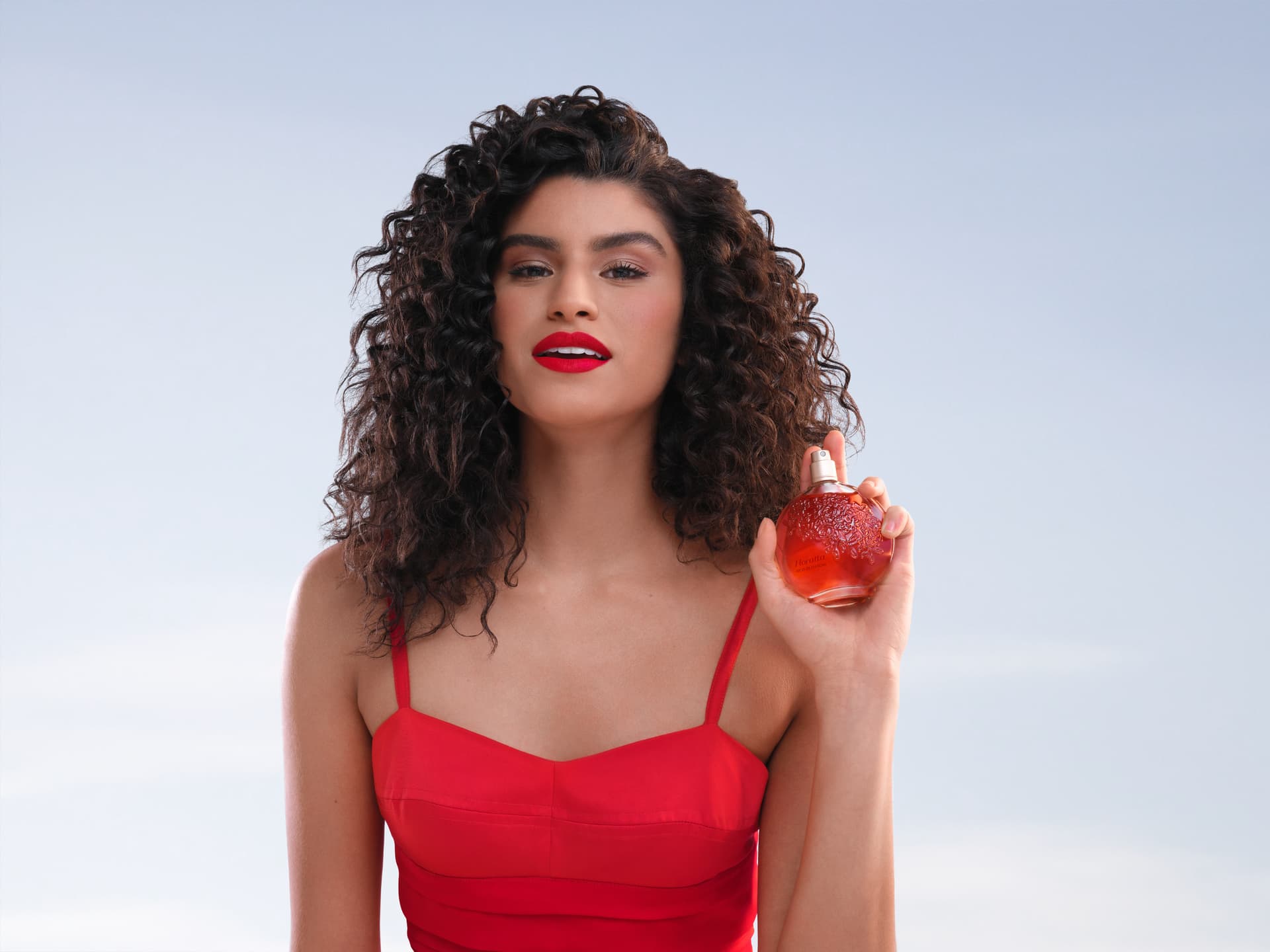 Imagem de mulher com cabelo cacheado, roupas e batom vermelho segurando o frasco da fragrância Floratta Red Blossom