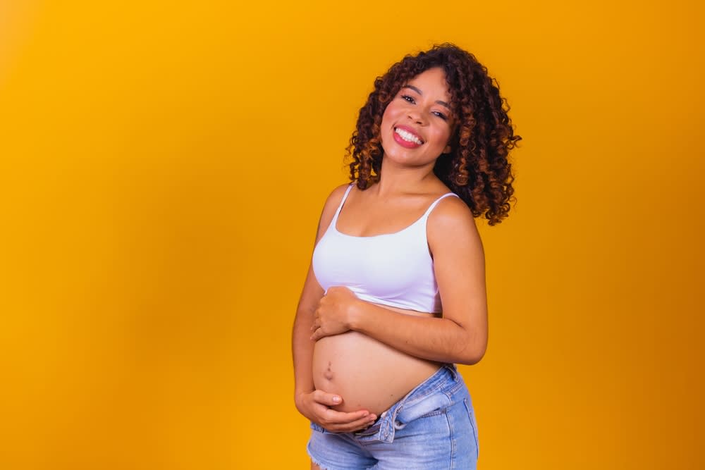 Imagem de mulher grávida, ela tem cabelos cacheados em tom escuro com luzes e está com a barriga de fora e suas mãos sobre elas