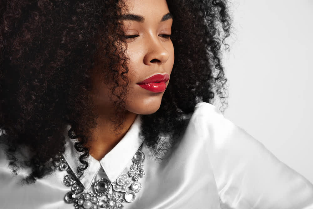 Imagem de mulher com blusa branca e cabelo cacheado na cor preta, ela está usando um batom vermelho com efeito vinil