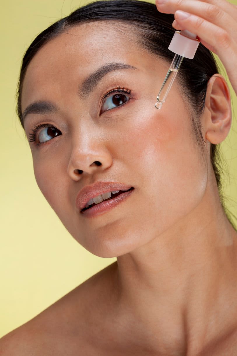Mulher de pele clara aplicando um sérum translúcido com um conta-gotas no lado de seu rosto, olhando para cima. O fundo é de um tom de amarelo suave.