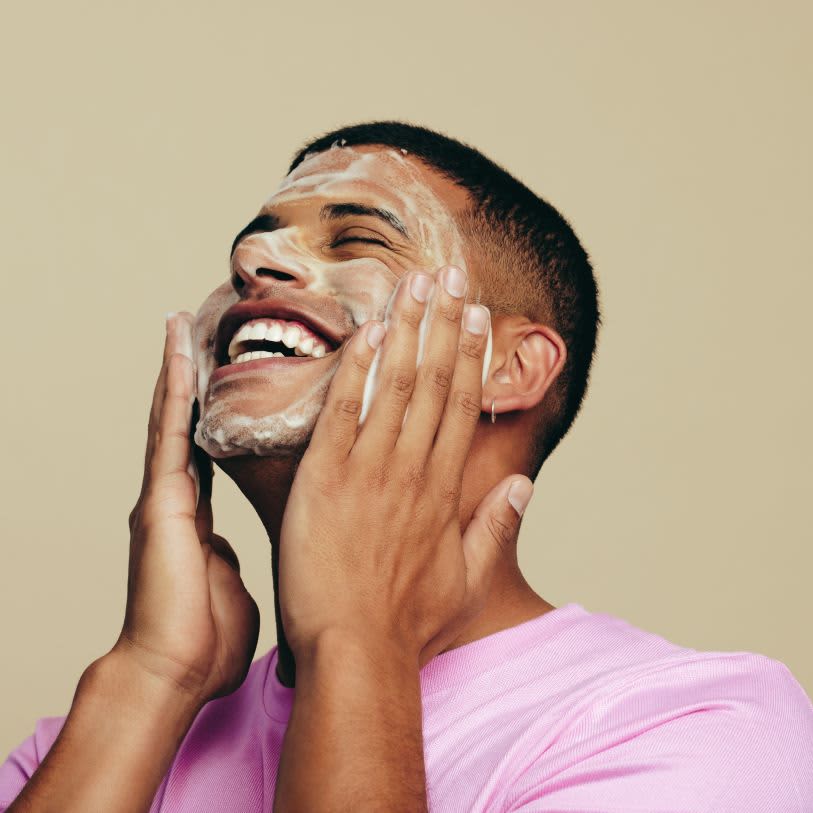 Homem feliz, de camiseta rosa, lavando o rosto com sabonete em espuma, massageando as bochechas, ideal para a limpeza da pele mista.