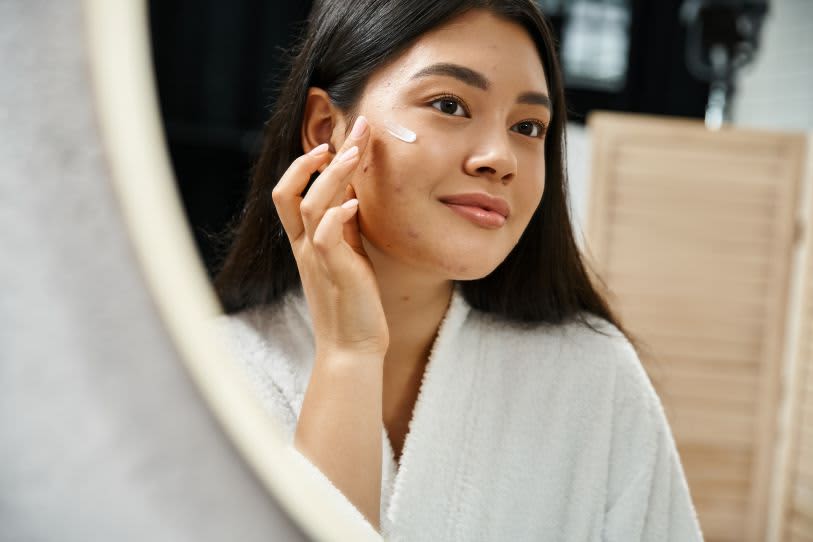 Mulher jovem com acne sorri ao aplicar creme facial no rosto, refletida em um espelho circular em um ambiente iluminado.
