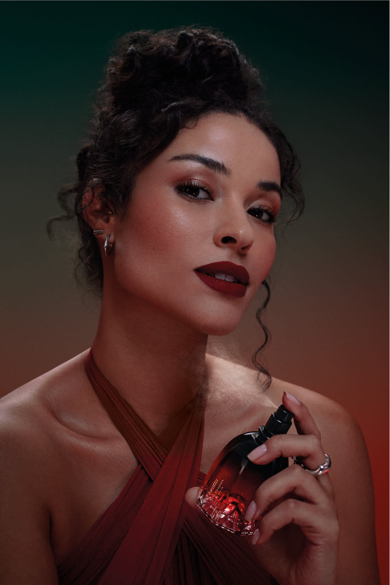 Uma mulher com cabelo cacheado e vestido vermelho está borrifando uma fragrância em seu pescoço. A marca do frasco é Floratta Red Passion.