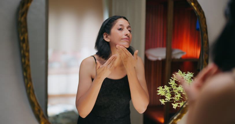 Mulher de pele branca e cabelo preto, liso e na altura dos ombros analisa a pele com um olhar de preocupação em frente ao espelho 