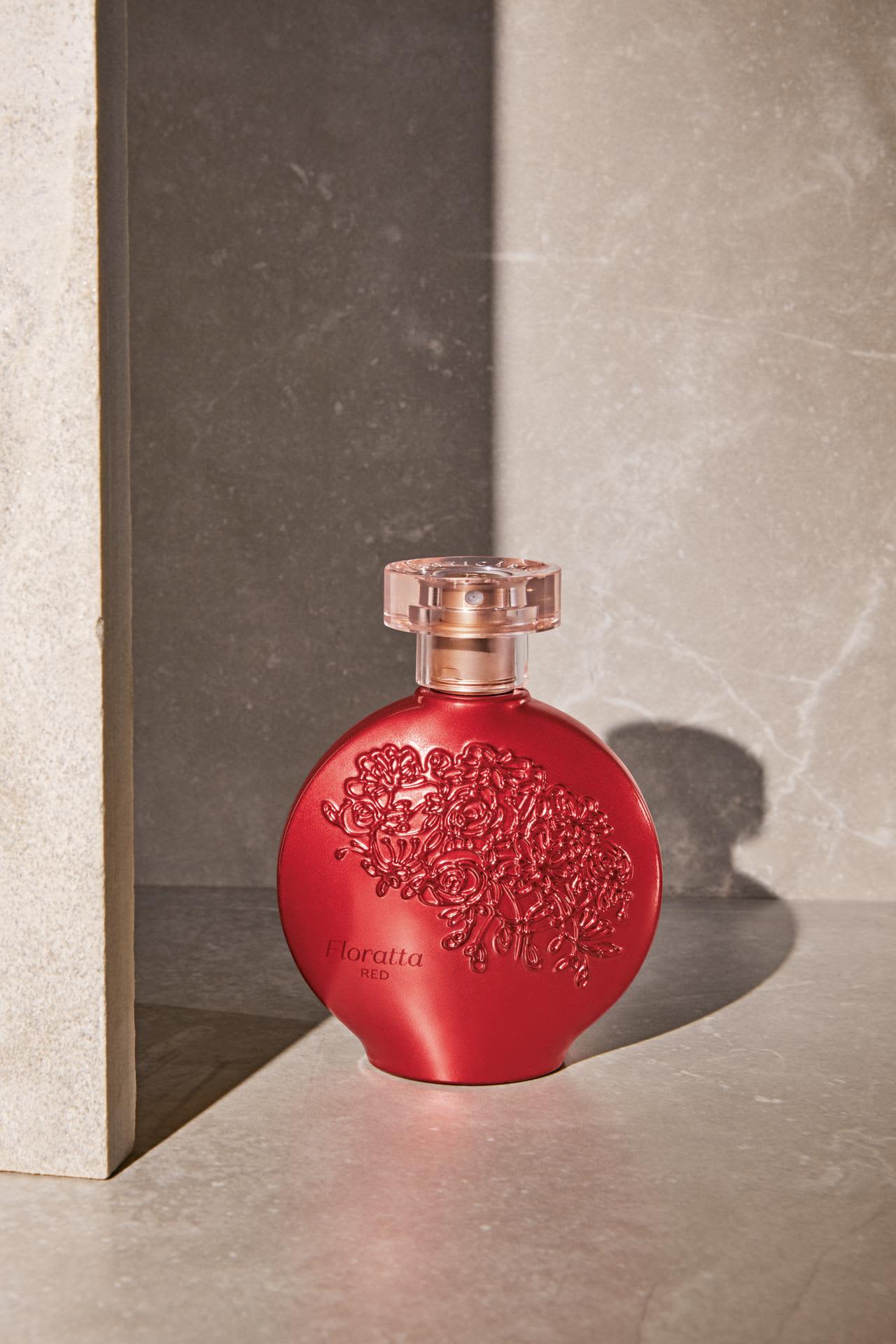 Frasco redondo da fragrância Floratta Red na cor vermelho metálico brilhante, com detalhes florais em relevo e tampa transparente. O frasco está posicionado em uma superfície de mármore cinza claro, entre dois painéis de mármore cinza escuro, com iluminação dramática criando sombras fortes.