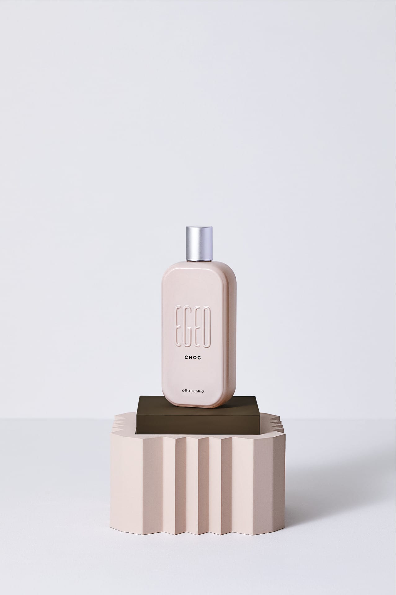 Frasco retangular da fragrância Egeo Choc em tom nude/rosa pálido com a palavra "EGEO" em relevo. O frasco está apoiado em um pequeno bloco marrom escuro que, por sua vez, está sobre um pedestal geométrico de cor creme claro. O fundo é branco e liso.