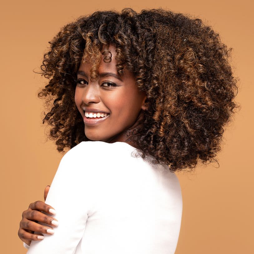 Uma mulher negra sorri para a câmera com os braços cruzados, vestindo uma blusa branca. Ela tem cabelos cacheados volumosos, com mechas em tons de castanho e loiro. O fundo da foto é de cor bege.