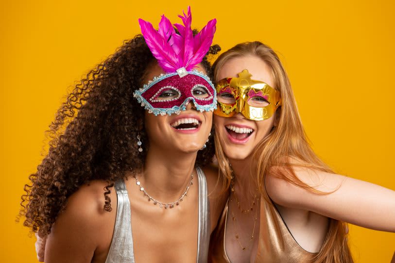Duas mulheres sorridentes vestindo máscaras coloridas de carnaval, uma com penas rosas e outra dourada, em um fundo amarelo vibrante. Elas estão abraçadas, transmitindo alegria e descontração.