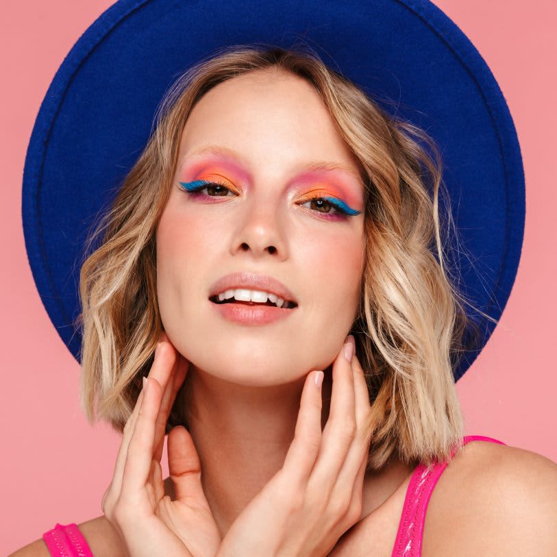 Mulher loira de chapéu azul e fundo rosa. Maquiagem artística com sombras rosa e laranja e delineado azul vibrante.