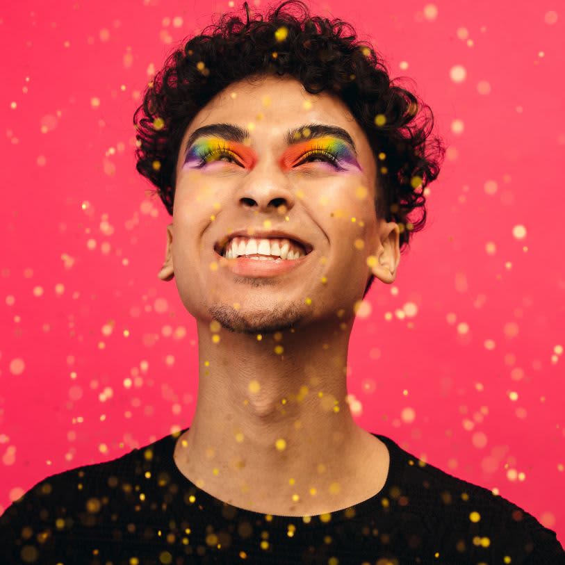 Homem jovem sorrindo sob confetes dourados. Ele usa sombra arco-íris multicolorida e está em um fundo rosa intenso.