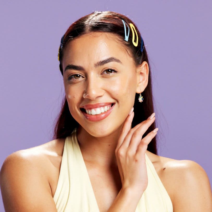 Close de mulher jovem sorrindo com pele iluminada e viçosa. Ela usa presilhas coloridas no cabelo e tem um fundo roxo suave.