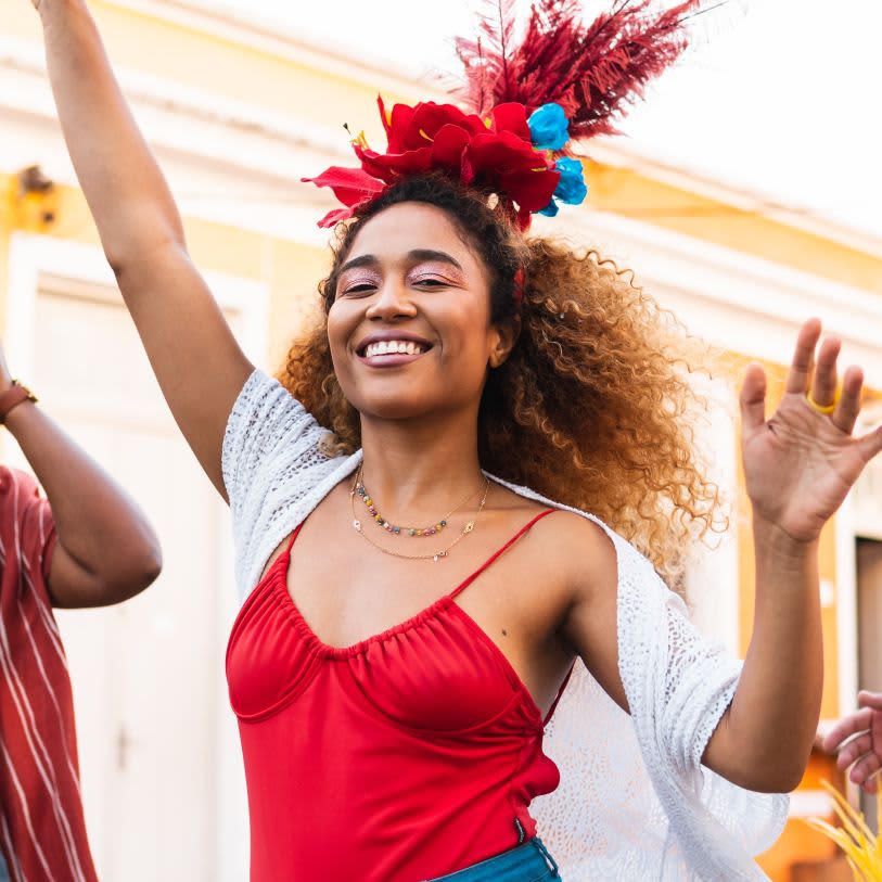 Mulher de cabelos cacheados celebrando com um adereço de flores vermelhas na cabeça. Ela usa sombra rosa cintilante e veste vermelho