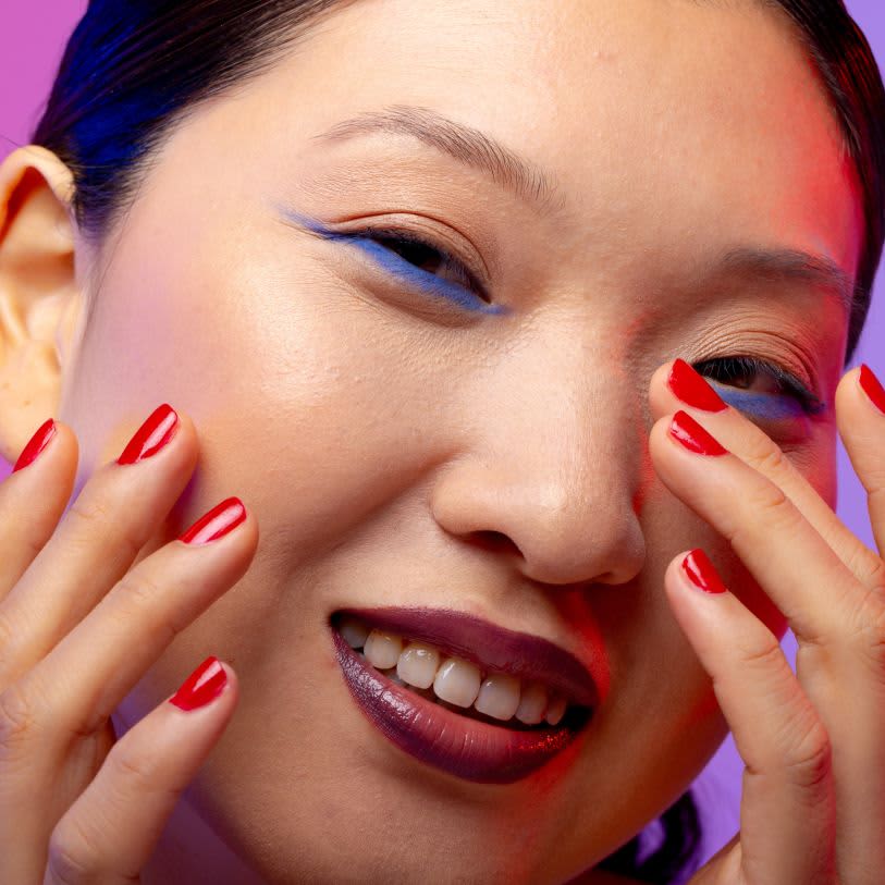 Close de rosto feminino sorrindo com delineado azul sutil, unhas vermelhas e batom roxo degradê. Fundo roxo e rosa.