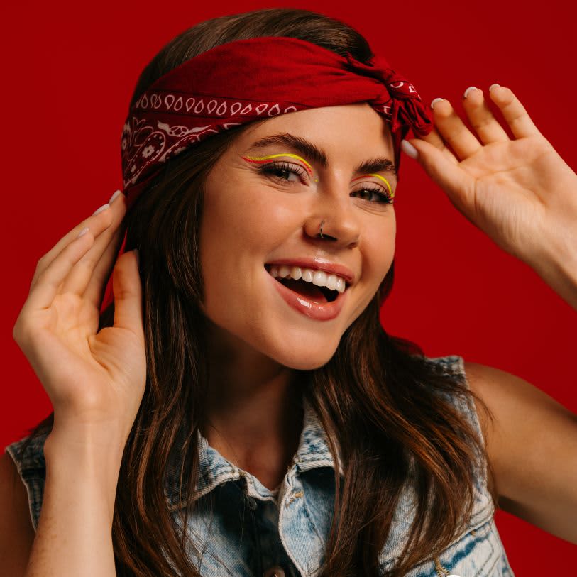 Mulher de bandana vermelha sorrindo. Ela usa delineado duplo gráfico nas cores amarelo e vermelho. Fundo vermelho sólido