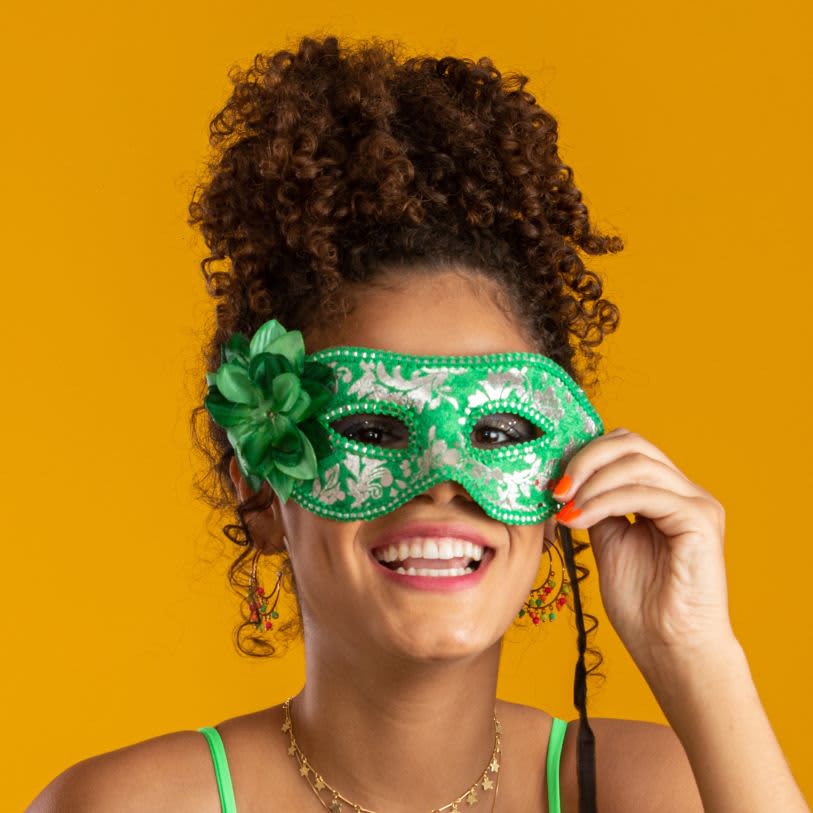 Mulher sorridente de cabelos cacheados presos em um coque abacaxi alto, usando máscara verde de Carnaval e acessório de flor.