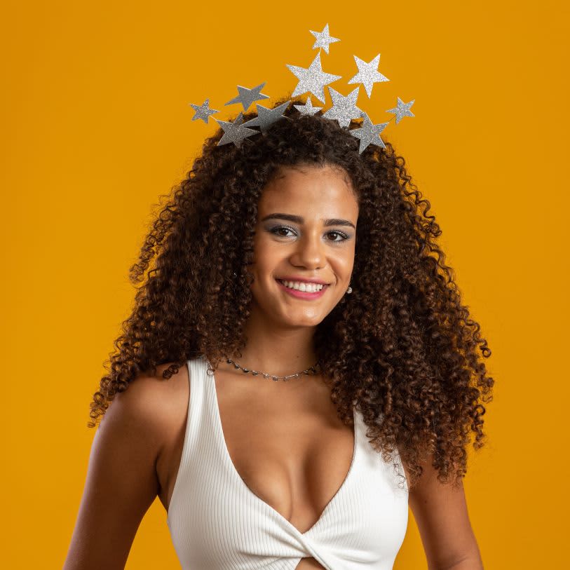 Mulher de cabelos cacheados soltos usando uma tiara de estrelas prateadas e blusa branca, sorrindo contra um fundo amarelo.