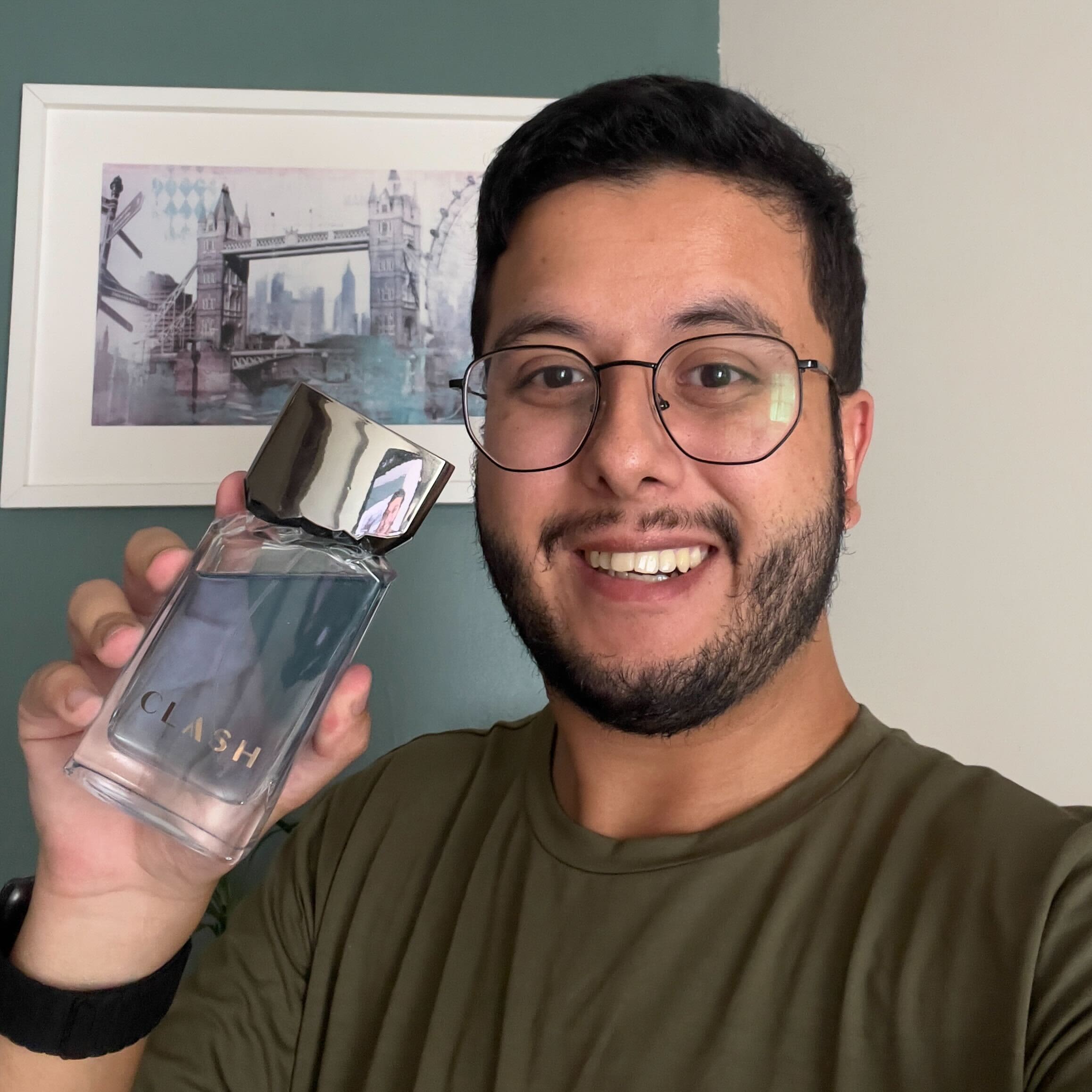 Homem branco com barba e cabelo preto sorrindo e segurando nas mãos o frasco de Clash