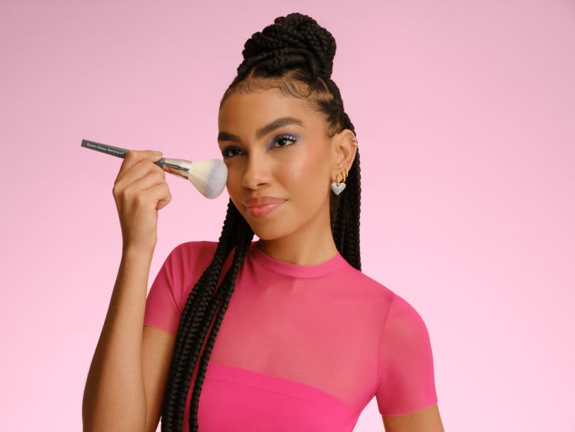 Uma jovem com tranças aplica pó com um pincel grande e macio. Ela está sorrindo para a câmera.