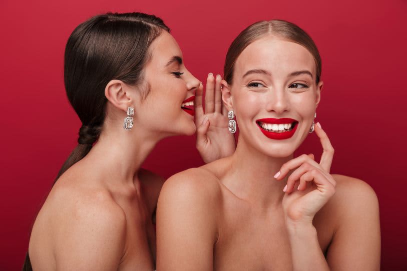 Duas mulheres elegantes com batom vermelho vibrante compartilham um segredo divertido, criando um clima de cumplicidade e glamour.
