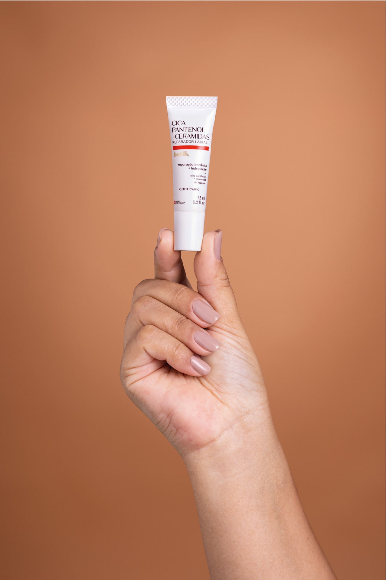 Uma mão segurando o produto Botik Cica Pantenol + Ceramidas, um reparador labial. Fundo marrom claro.