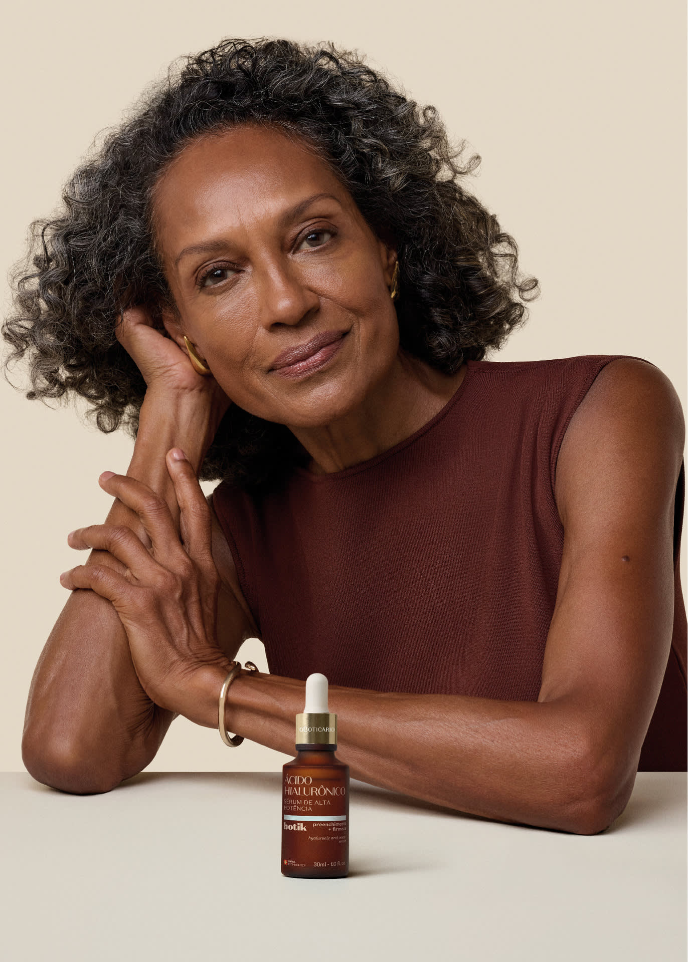 Mulher com cabelos cacheados escuros sorri levemente, apoiando a cabeça na mão, ao lado de um frasco do Sérum de Alta Potência Ácido Hialurônico Botik