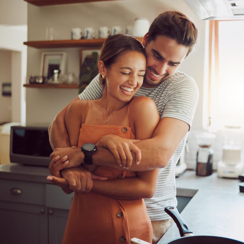 Homem abraça mulher por trás em uma cozinha; ambos sorriem felizes e demonstram afeto
