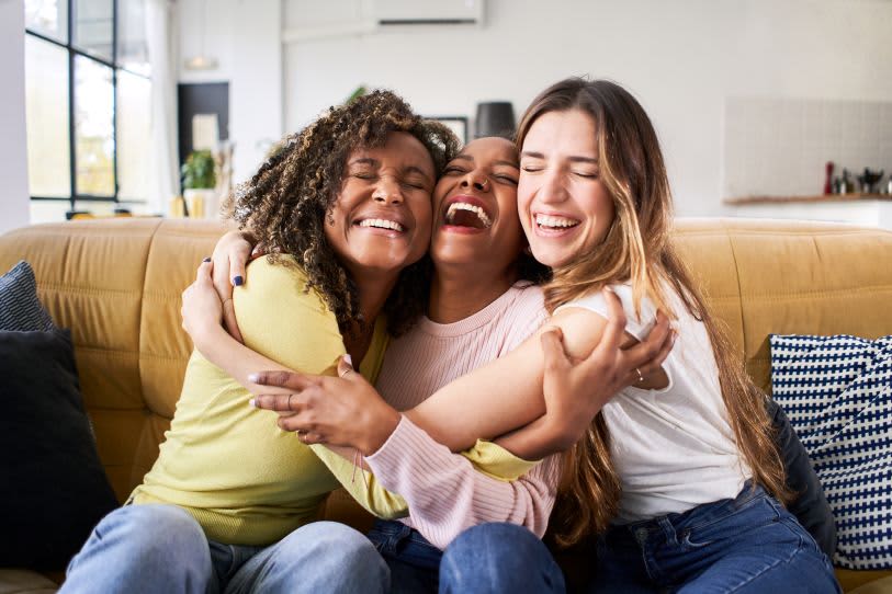 Três amigas sorriem e se abraçam alegremente em um sofá, celebrando a amizade em um ambiente iluminado e acolhedor.