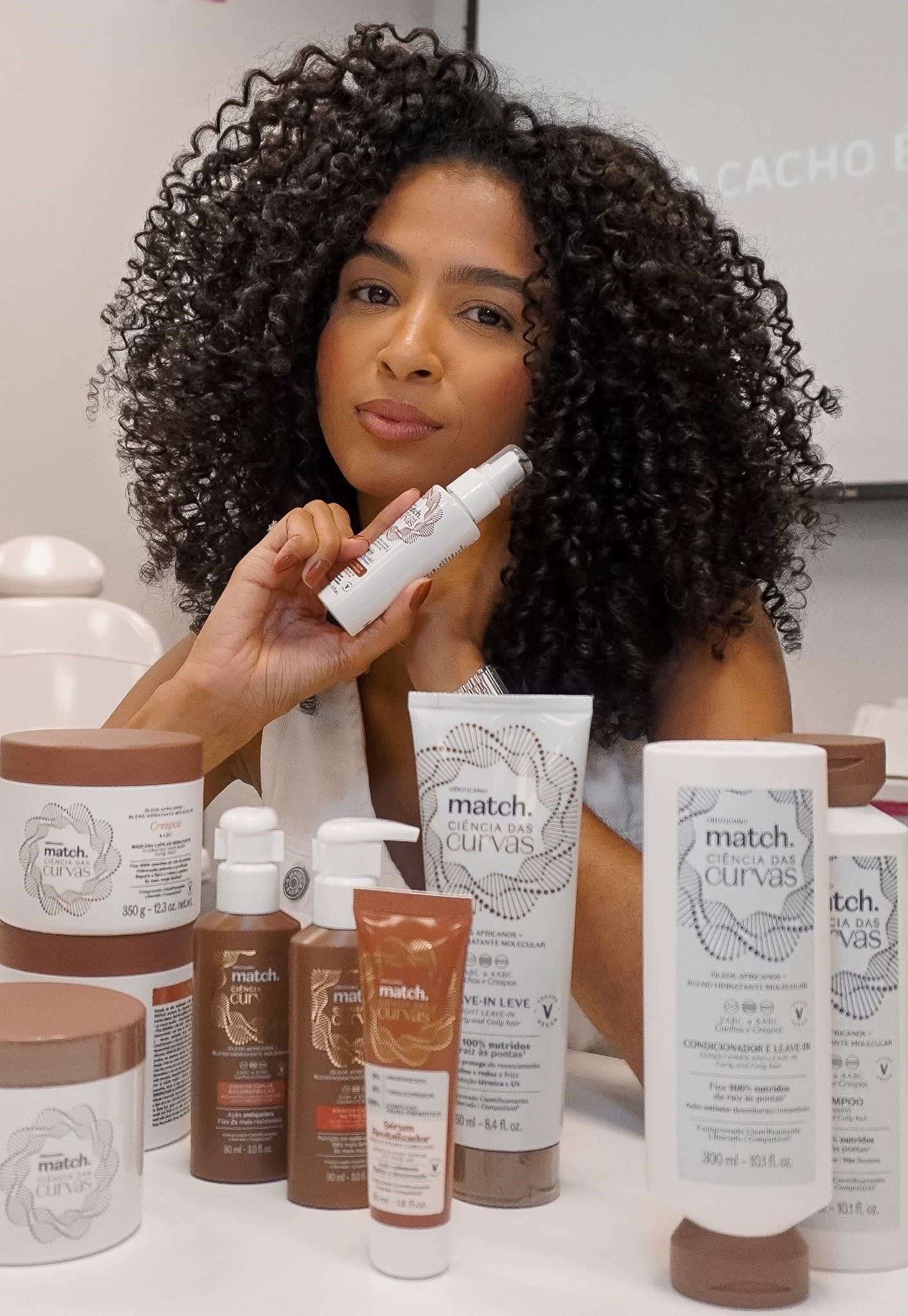 Produtos para cabelos cacheados em cima de uma mesa branca e atrás a influenciadora Thuany Cordeiro