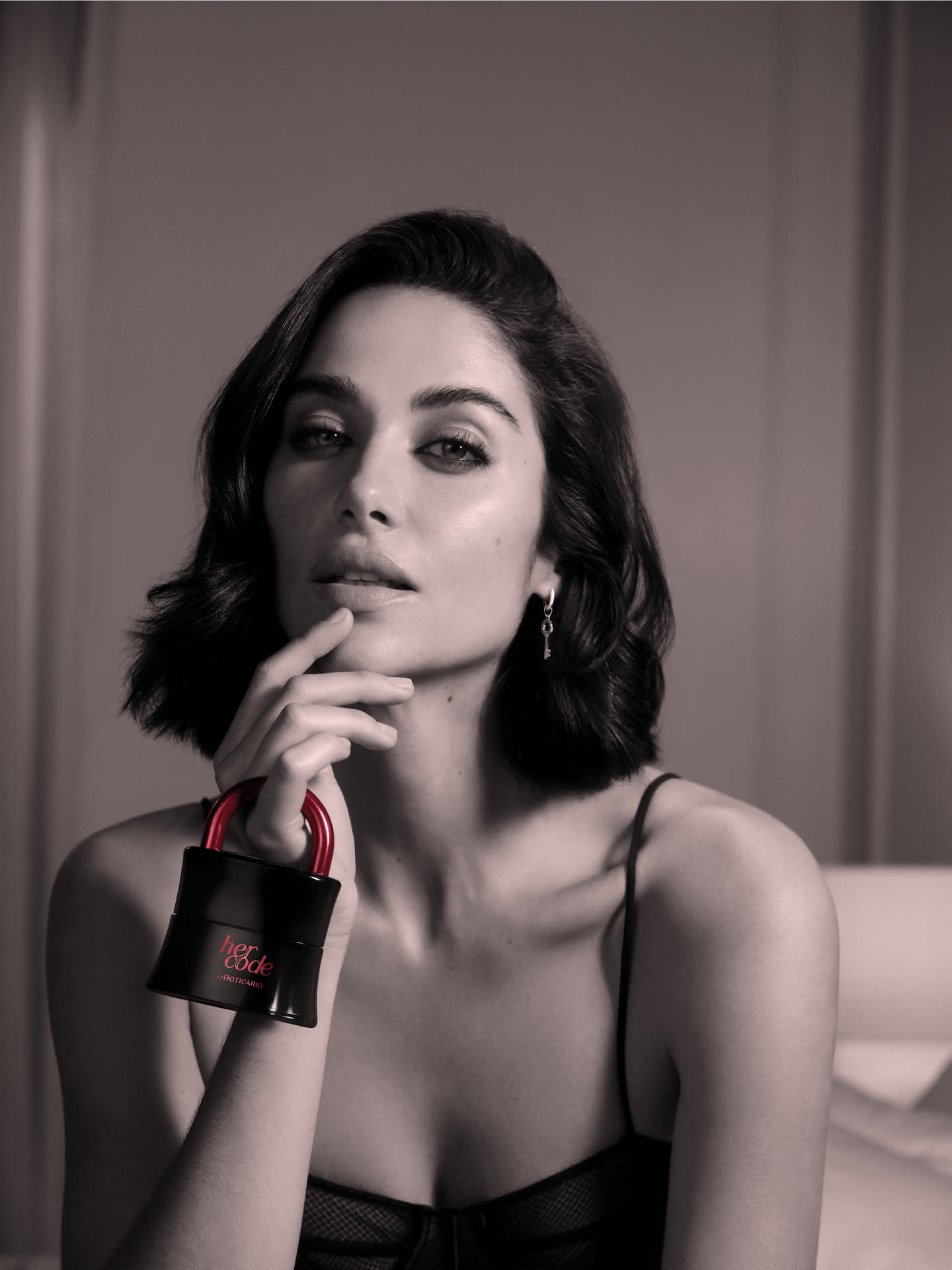 Close-up de uma mulher com cabelo escuro segurando um frasco de fragrância em forma de cadeado, olhando para a câmera.