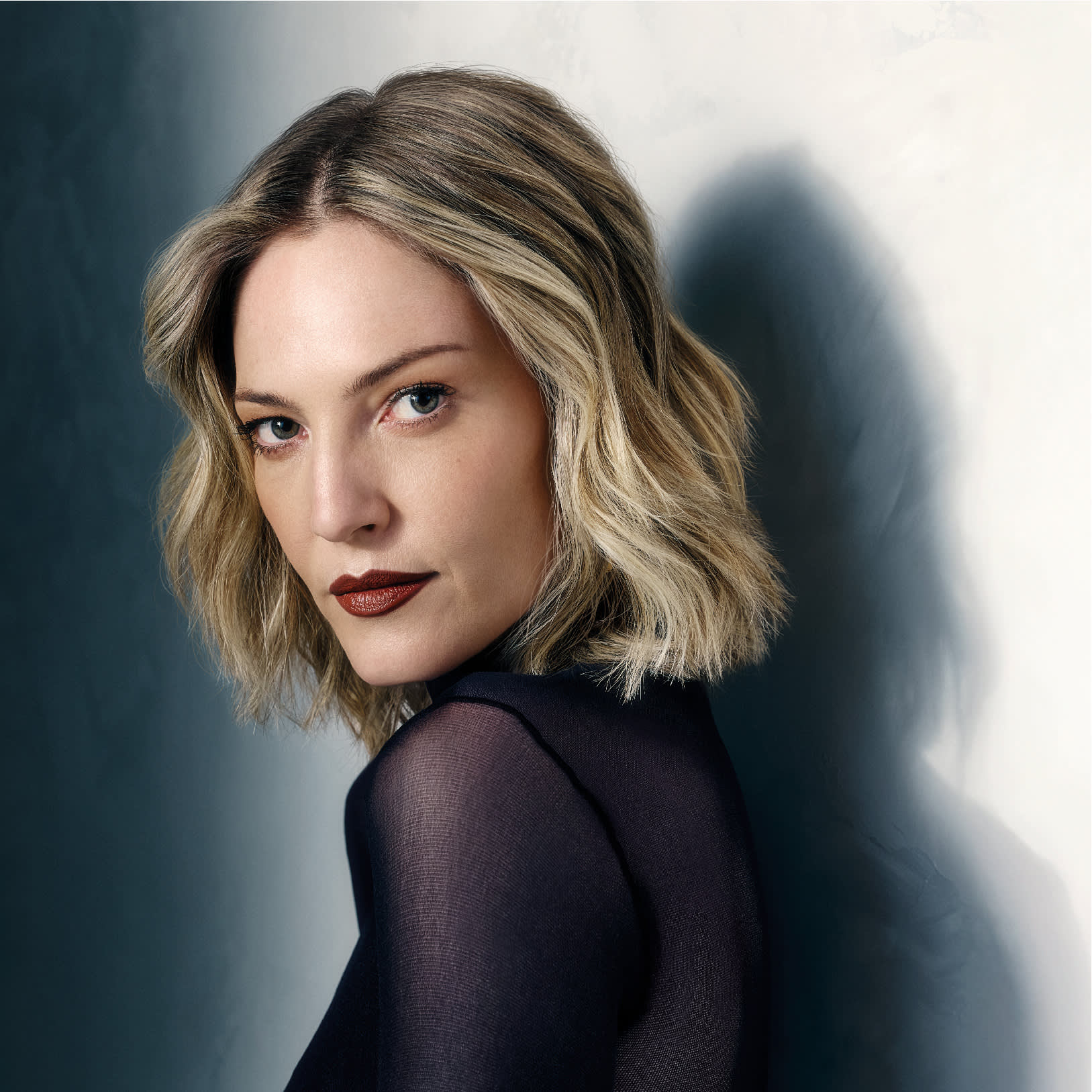 Uma mulher com cabelo loiro na altura dos ombros e batom escuro olha por cima do ombro