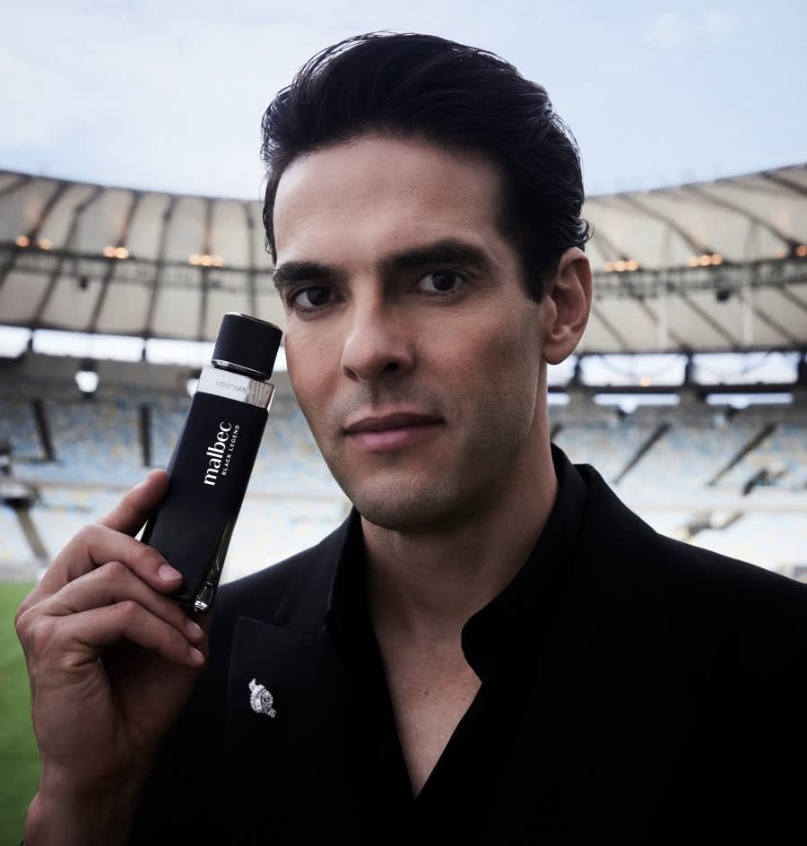 Kaká segura a fragrância Malbec Black Legend próximo ao rosto. Ele veste uma roupa preta e está no Estádio do Maracanã, no Rio de Janeiro