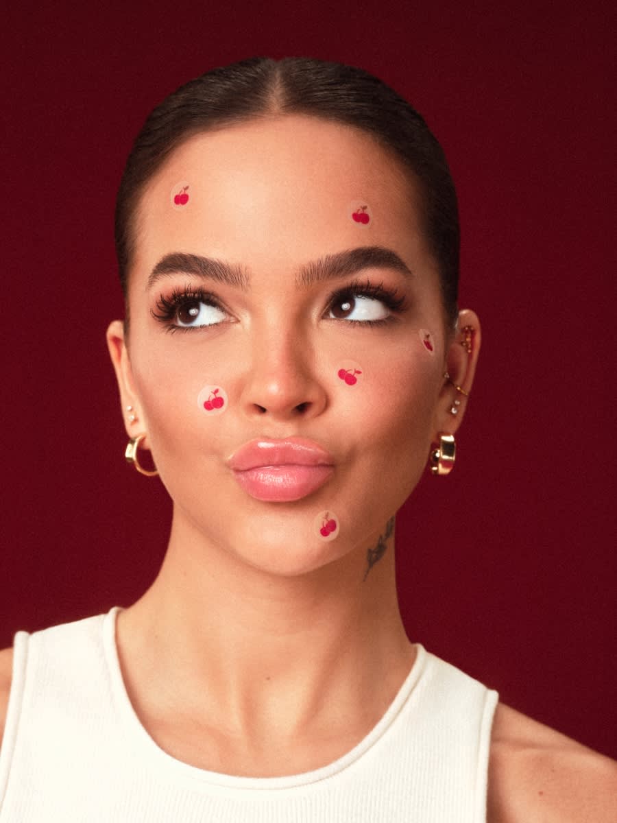 A atriz Mel Maia veste uma blusa clara, está de cabelo preto, com brincos dourados e com adesivos em formato de cereja espalhados pelo rosto. O fundo é na cor vinho.