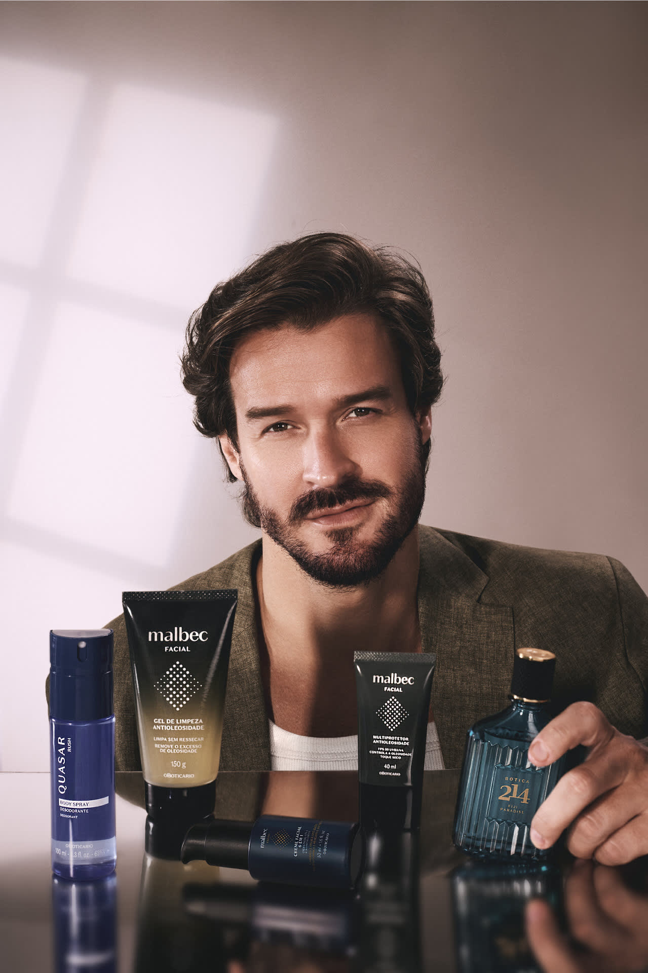 Homem sorridente com produtos Malbec Facial, Quasar e Botica 214 sobre base refletora.