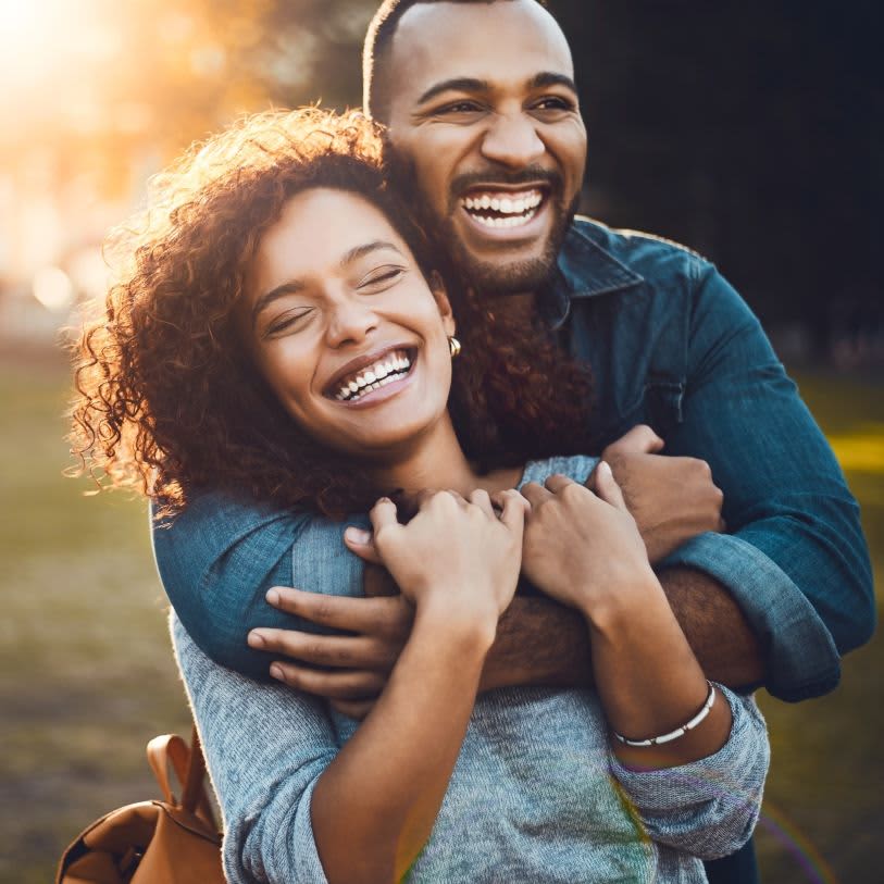 Homem negro abraça mulher negra; ambos sorriem alegremente em um parque ensolarado.