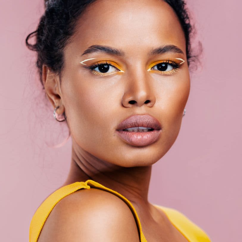 Uma mulher negra exibe uma maquiagem artística marcante com sombra amarela vibrante e delineado branco vazado, vestindo roupa amarela sobre um fundo rosa.