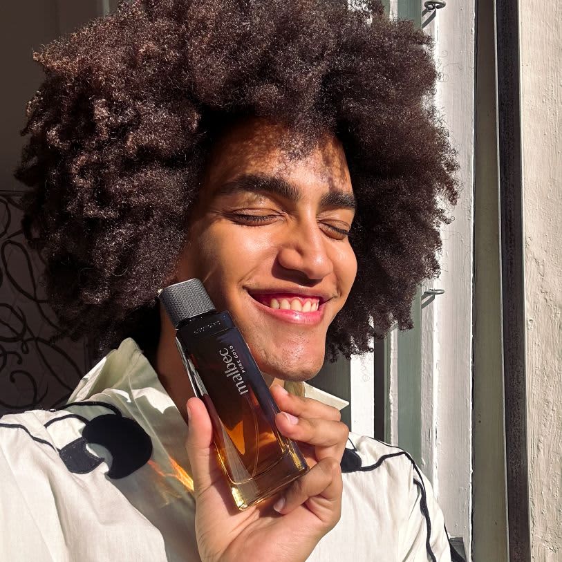 Homem sorridente com cabelo crespo segura um frasco da fragrância Malbec Pure Gold sob a luz do sol, em uma composição calorosa e iluminada.