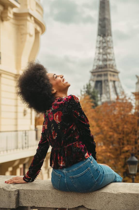 Imagem de mulher sorrindente sentada. Ao fundo está a Torre Eiffel