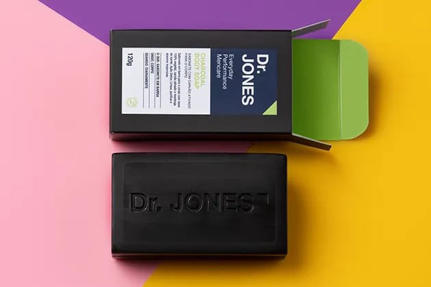 Sabonete de Carvão Vegetal Charcoal Body Soap, da Dr. JONES em fundo rosa, roxo e amarelo.