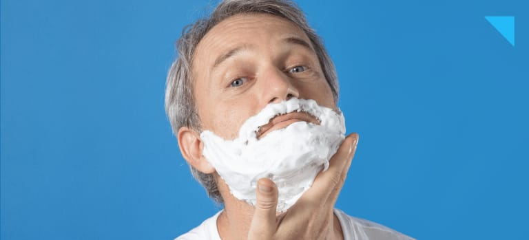 Em fundo azul, homem de cabelos grisalhos, pele clara e camiseta branca passa a espuma de barbear Precision Foam, da Dr. JONES, na sua barba.