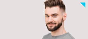 Foto de homem de camisa cinza em fundo neutro, cabelo raspado na lateral, meio sorriso e barba degradê.