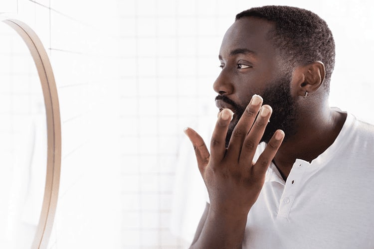 Homem de pele preta se olhando no espelho enquanto passa a mão pela sua barba.