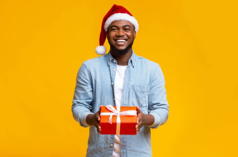 Homem com chapéu vermelho de papai noel, vestindo uma camisa azul clara por cima de uma camiseta branca, segurando uma caixa de presente vermelha com laço branco.