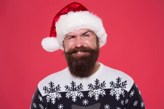 Homem com gorro vermelho e branco de Natal, com barba longa e castanha, vestindo uma blusa de lã com flocos de neve. Fundo vermelho.
