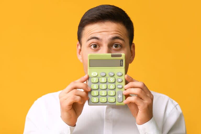 Homem segurando uma calculadora, que cobre quase todo o seu rosto, apenas com os olhos em expressão de surpresa. Representando o assunto "vida financeira".