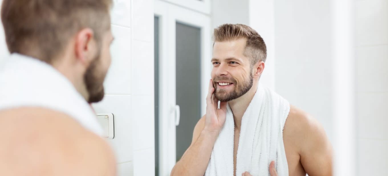 Homem sem camisa com toalha branca pendurada em seu pescoço, observando seu reflexo no espelho do banheiro enquanto passa a mão sobre sua barba e sorri.