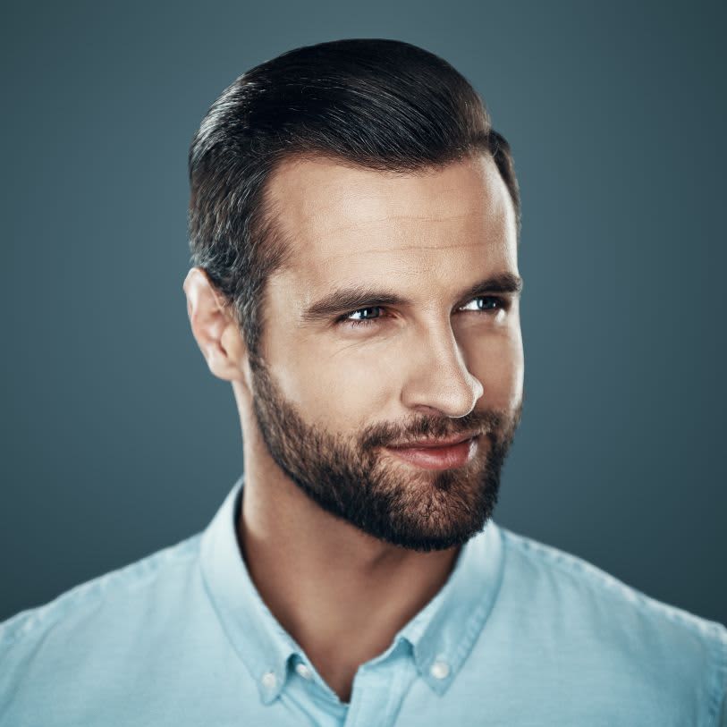 Homem com barba bem alinhada e cabelo penteado para trás, vestindo uma camisa azul clara. Ele tem um olhar confiante e um leve sorriso, posando contra um fundo neutro.