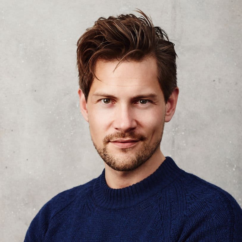 Homem branco de cabelos castanhos usando um suéter azul-marinho. Ele está olhando para a câmera com uma expressão neutra