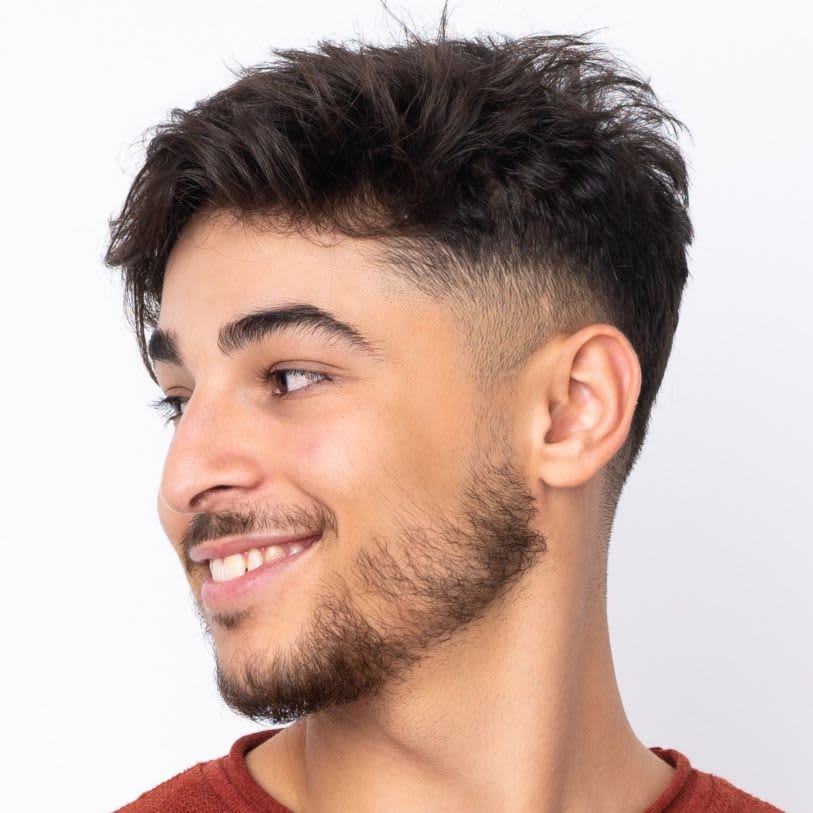 Jovem sorridente com corte americano masculino. Ele olha para a direita, sobre um fundo branco.